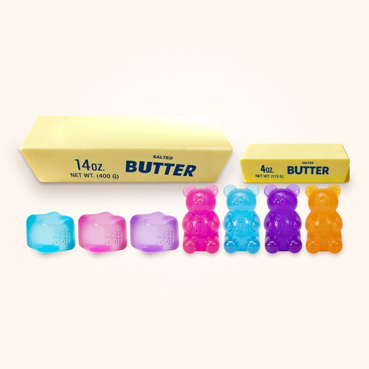 Squishy Gift Box Everything: all three NeeDoh Nice Cube colors and all four NeeDoh Gummy Bear colors in front of a Giant and a Mini Butter Squishy, shown to scale on a cream background