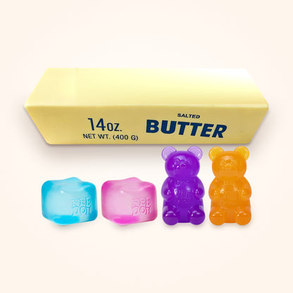 Squishy Gift Box Family: blue and pink NeeDoh Nice Cubes and purple and orange NeeDoh Gummy Bears in front of a Giant Butter Squishy, shown to scale on a cream background